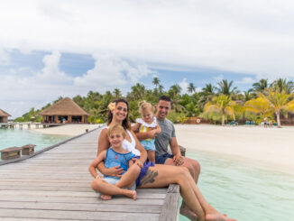 一家四口坐在木制码头上，面带微笑，背景是碧绿的海水和热带沙滩。.