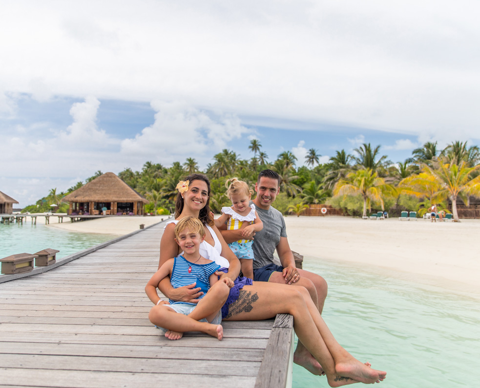 Una familia de cuatro personas está sentada en un muelle de madera, sonriendo, con aguas turquesas y una playa tropical de fondo.