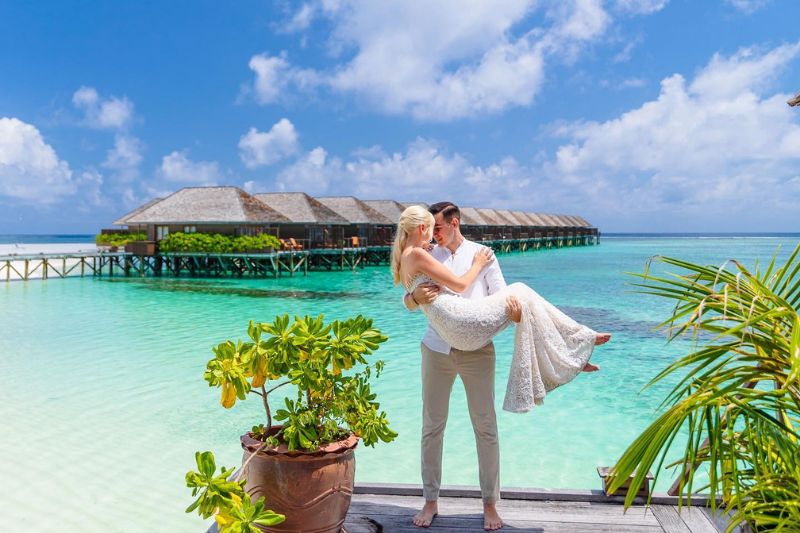 一对新人站在碧绿的海水边的木制码头上，男子抱着身穿白色婚纱的新娘，背景是热带风情的水上别墅。.