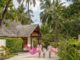 Family walks along a tropical resort path toward a thatched-roof building, each carrying large pink inflatable flamingos among palm trees and lush greenery.