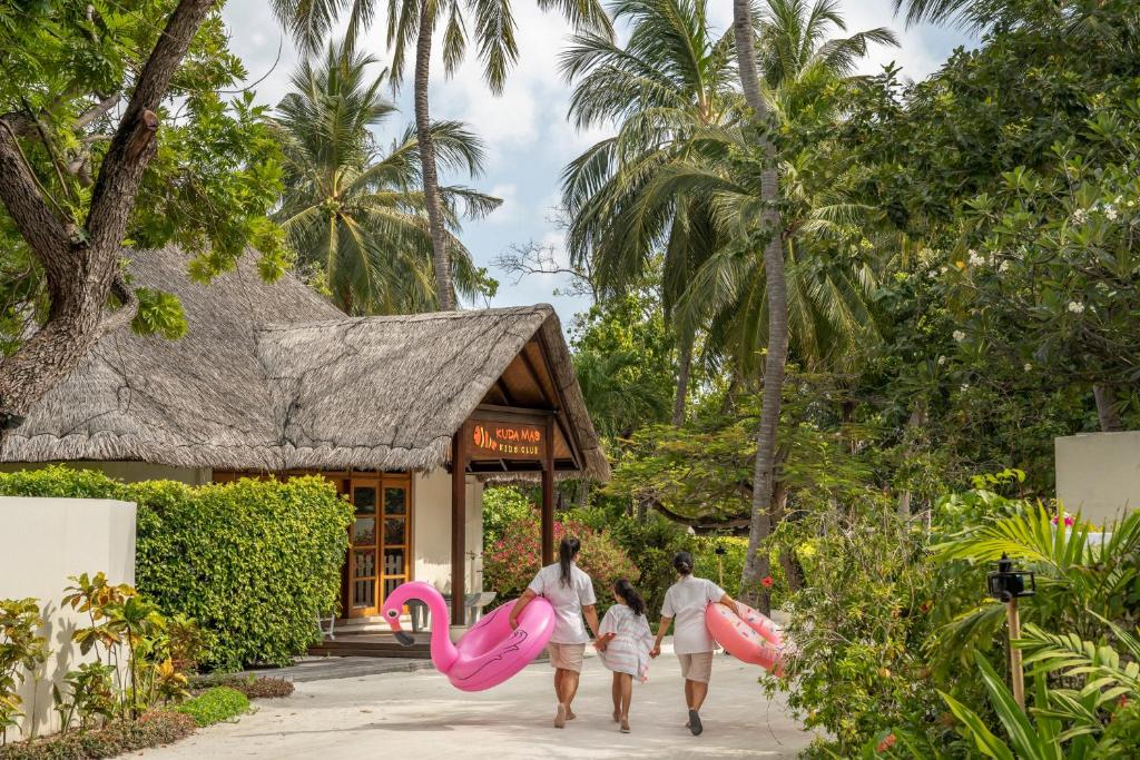 Una familia camina por un sendero de un complejo turístico tropical hacia un edificio con techo de paja, cada uno portando grandes flamencos inflables de color rosa entre palmeras y exuberante vegetación.