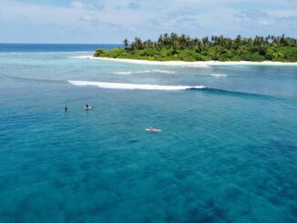A tropical island with a white-sand beach and palm trees, turquoise water, two surfers in the foreground, and a small boat nearby.
