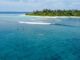 Une île tropicale avec une plage de sable blanc et des palmiers, une eau turquoise, deux surfeurs au premier plan et un petit bateau à proximité.