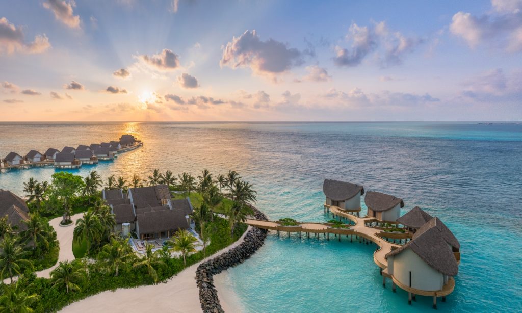 Vista aérea de un complejo turístico tropical con bungalows sobre el agua con techo de paja, conectados por una pasarela curva sobre aguas turquesas al atardecer.