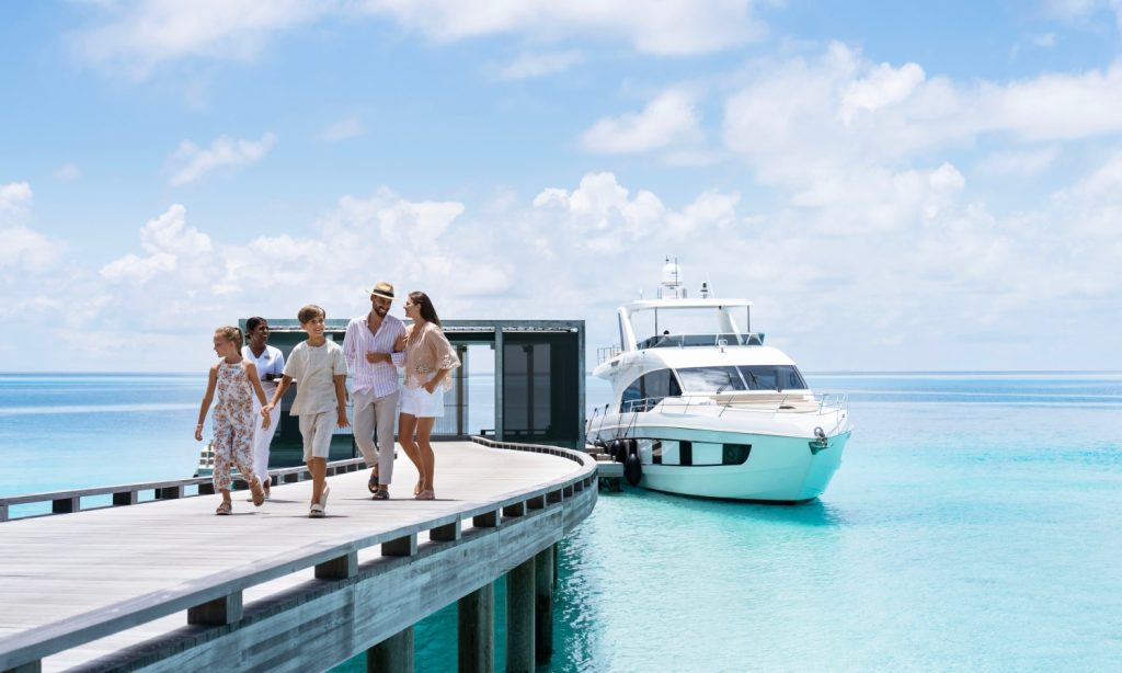 六个人沿着木制码头走向停泊在碧绿大海中的白色游艇，天空多云。.