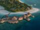 Aerial view of a tropical island resort with thatched overwater bungalows connected by boardwalks to a white-sand beach and turquoise ocean.