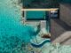 Aerial view of an overwater villa with a private rectangular pool, curved slide, wooden deck, and turquoise lagoon.