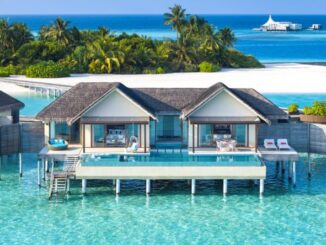 Overwater bungalow resort with thatched roofs on turquoise lagoon and palm trees in the background.