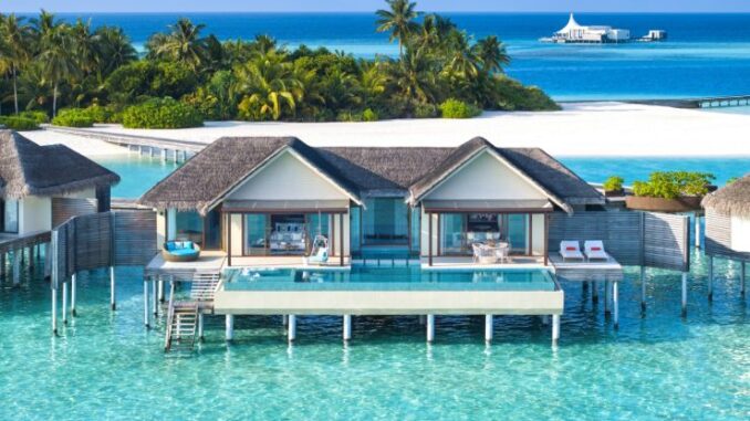 Overwater bungalow resort with thatched roofs on turquoise lagoon and palm trees in the background.