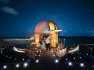 A couple stands inside a small domed observatory on a wooden deck by the ocean at night, under a starry sky with a telescope inside the dome near them.