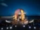 A couple stands inside a small domed observatory on a wooden deck by the ocean at night, under a starry sky with a telescope inside the dome near them.